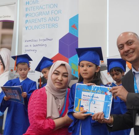 Three children are dressed in blue graduation caps and gowns. One child smiles at the camera holding a certificate with two adults beside her Image