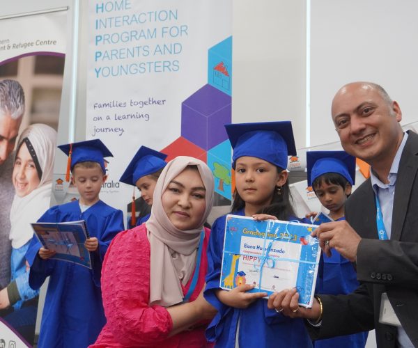 Three children are dressed in blue graduation caps and gowns. One child smiles at the camera holding a certificate with two adults beside her