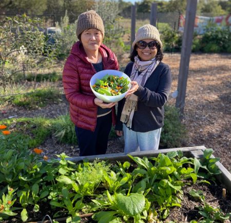 two women holding a bowl of salad. they are standing in front of a vegetable patch Image