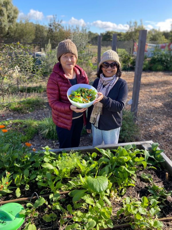 two women holding a bowl of salad. they are standing in front of a vegetable patch