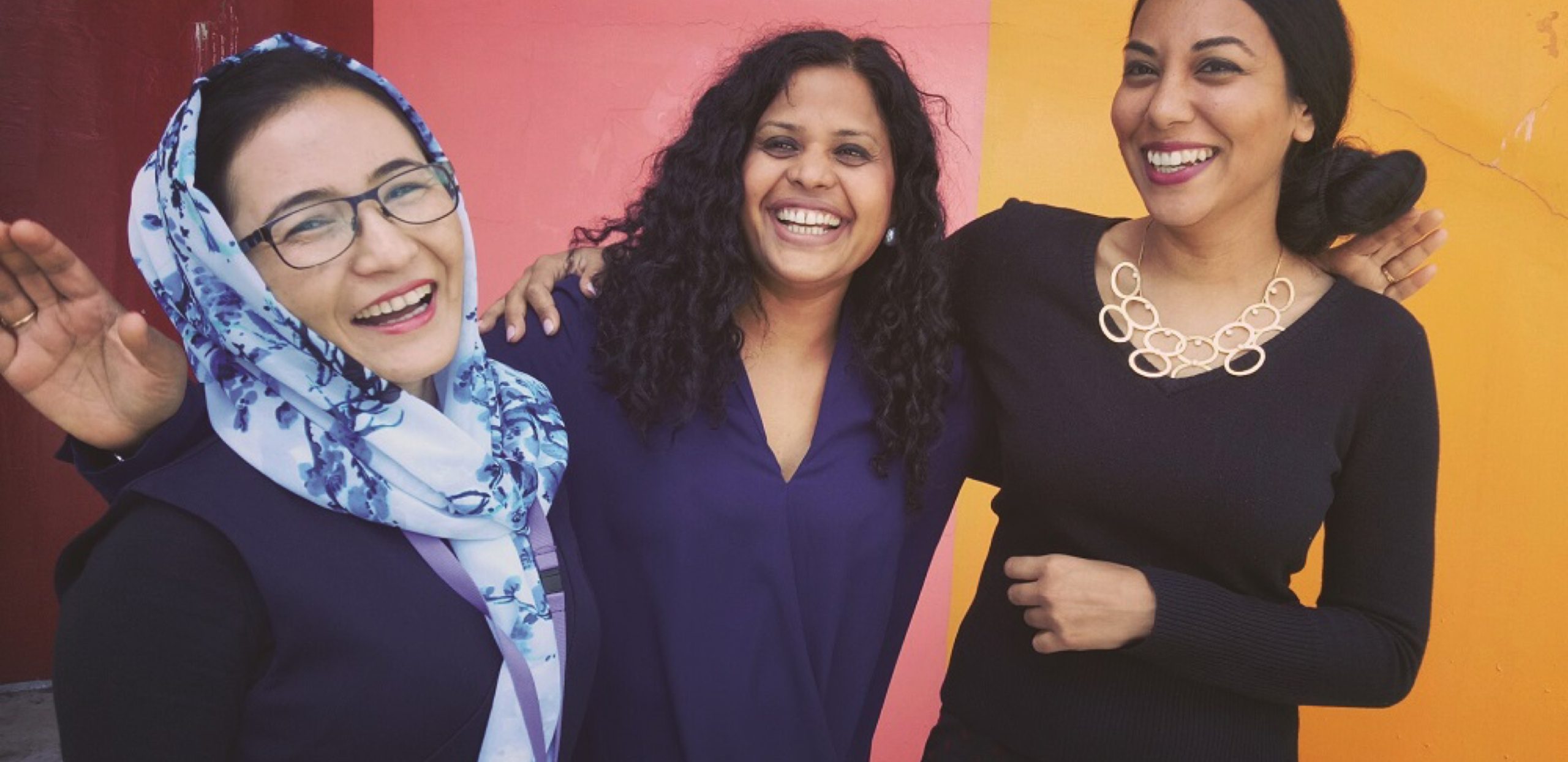 three women of different ethnic backgrounds laughing and smiling at camera in front of colourful wall