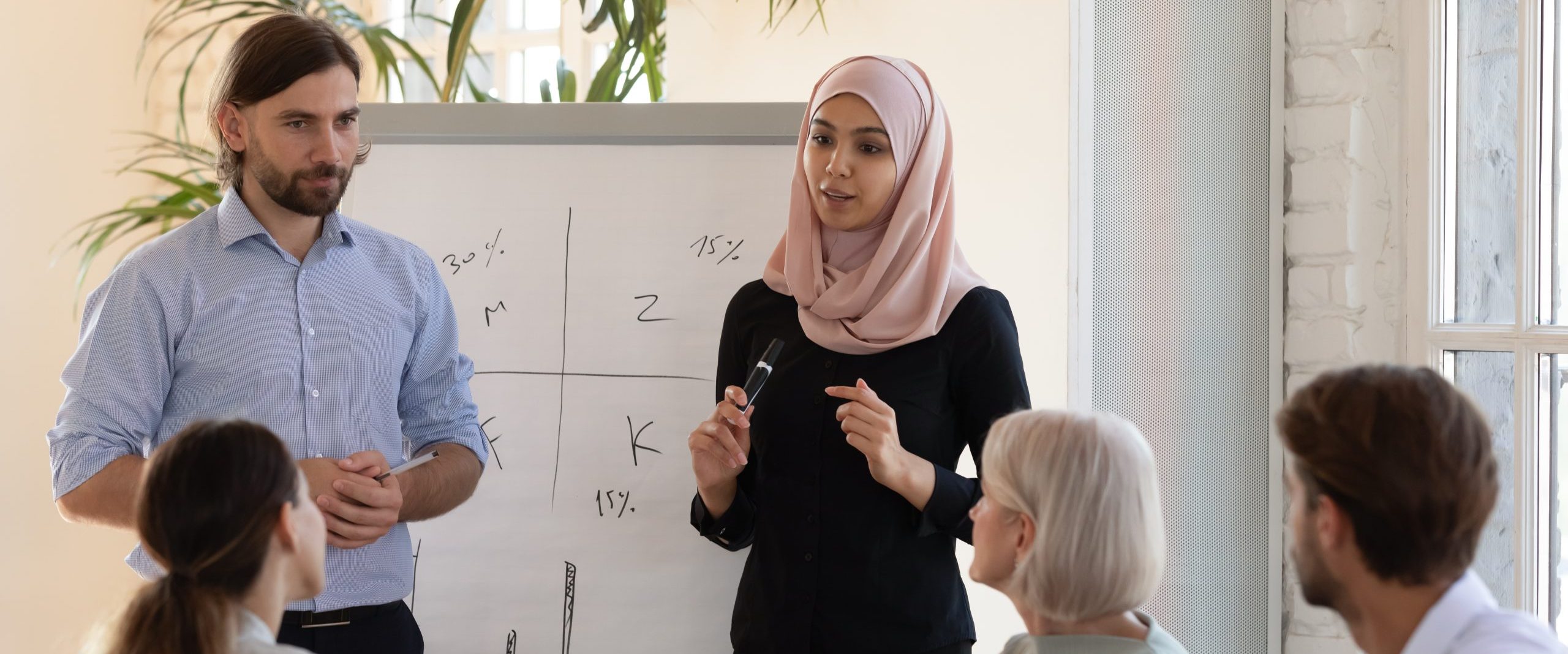 a woman in a headscarf and a man with a blue shirtpresenting to a room of three people