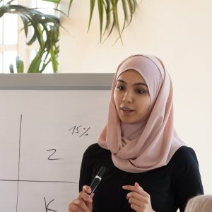 a woman in a headscarf and a man with a blue shirtpresenting to a room of three people