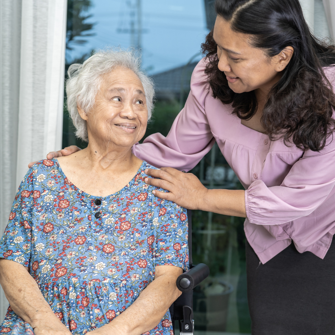 An older woman with grey hair is sitting and smiling up at a younger woman with dark hair