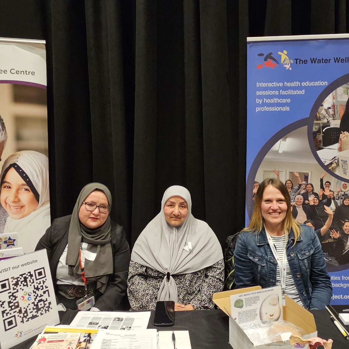 Three women sit behind a table with black cloth and flyers.