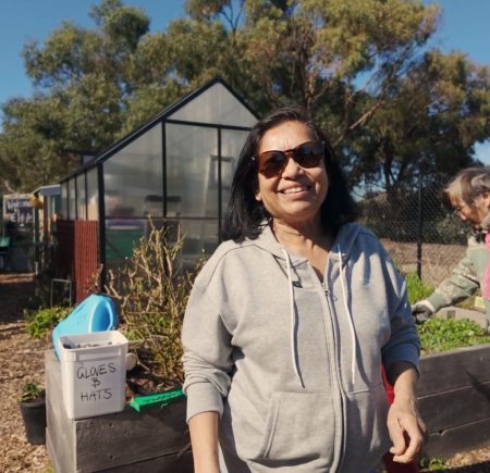 Shamalie smiles at the camera. She is wearing a pair of sunglasses and a woman is tending to a vegetable bed behind her.