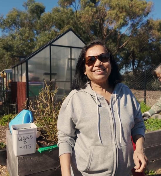 Shamalie smiles at the camera. She is wearing a pair of sunglasses and a woman is tending to a vegetable bed behind her.