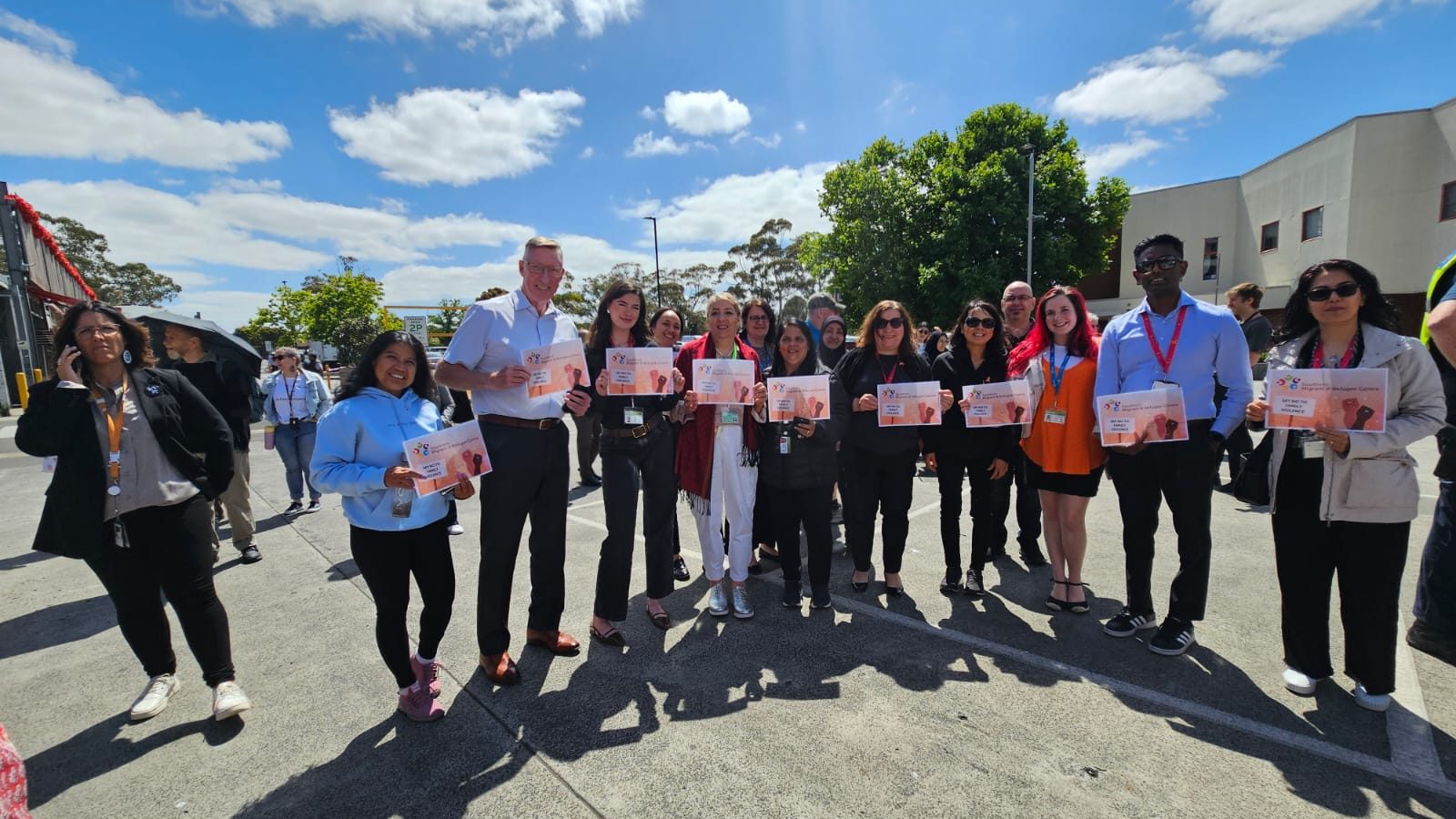 A group of SMRC community standing together outdoors, holding flyers and smiling towards the camera under a bright blue sky with scattered clouds. The diverse group includes men and women, some wearing name tags and lanyards.