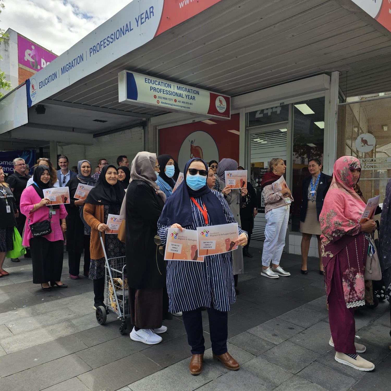 A diverse group of people standing outside a building. Many in the group are holding flyers, some wearing headscarves, and one individual is wearing a face mask.