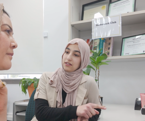 Samira (left) is looking at a computer screen while Arzo (right) points at the screen and speaks to Samira. They are both wearing light coloured head scarves.