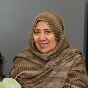 Two women standing and smiling. The woman on the right has dark hair and glasses. The woman on the right has a headscarf and is holding a sheet of green dough