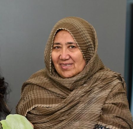 Two women standing and smiling. The woman on the right has dark hair and glasses. The woman on the right has a headscarf and is holding a sheet of green dough Image