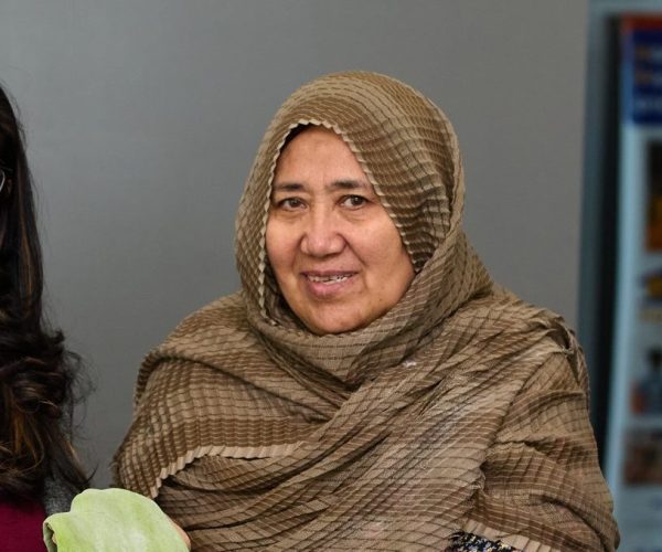 Two women standing and smiling. The woman on the right has dark hair and glasses. The woman on the right has a headscarf and is holding a sheet of green dough