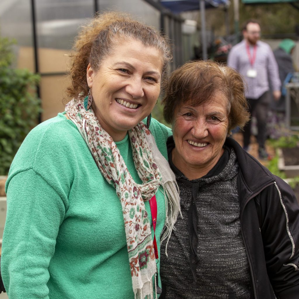Two women smile at the camera. They are standing in front of a community garden.