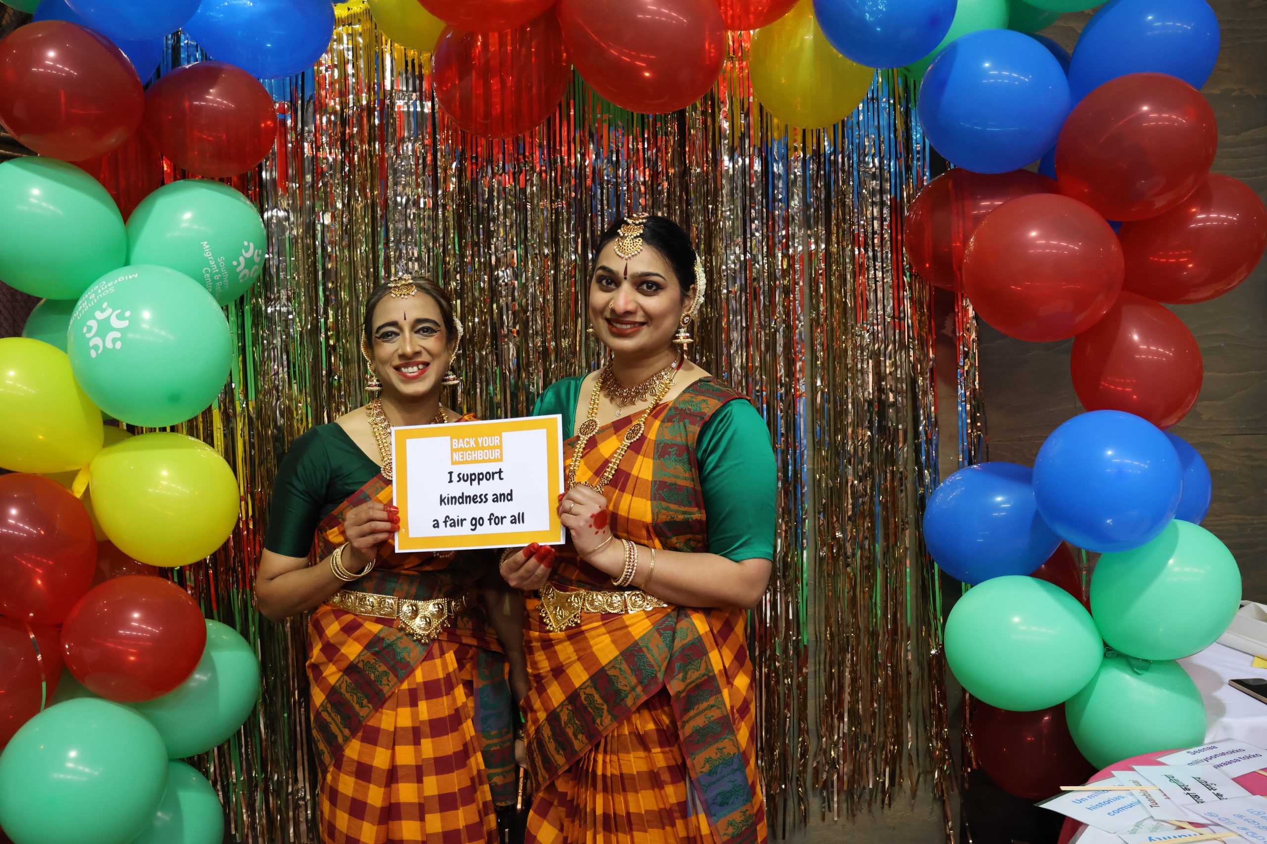 Two women dressed in Indian dress stand in front of a gold wall and colourful balloons. They hold a sign that says 'I support kindness and a fair go for all'