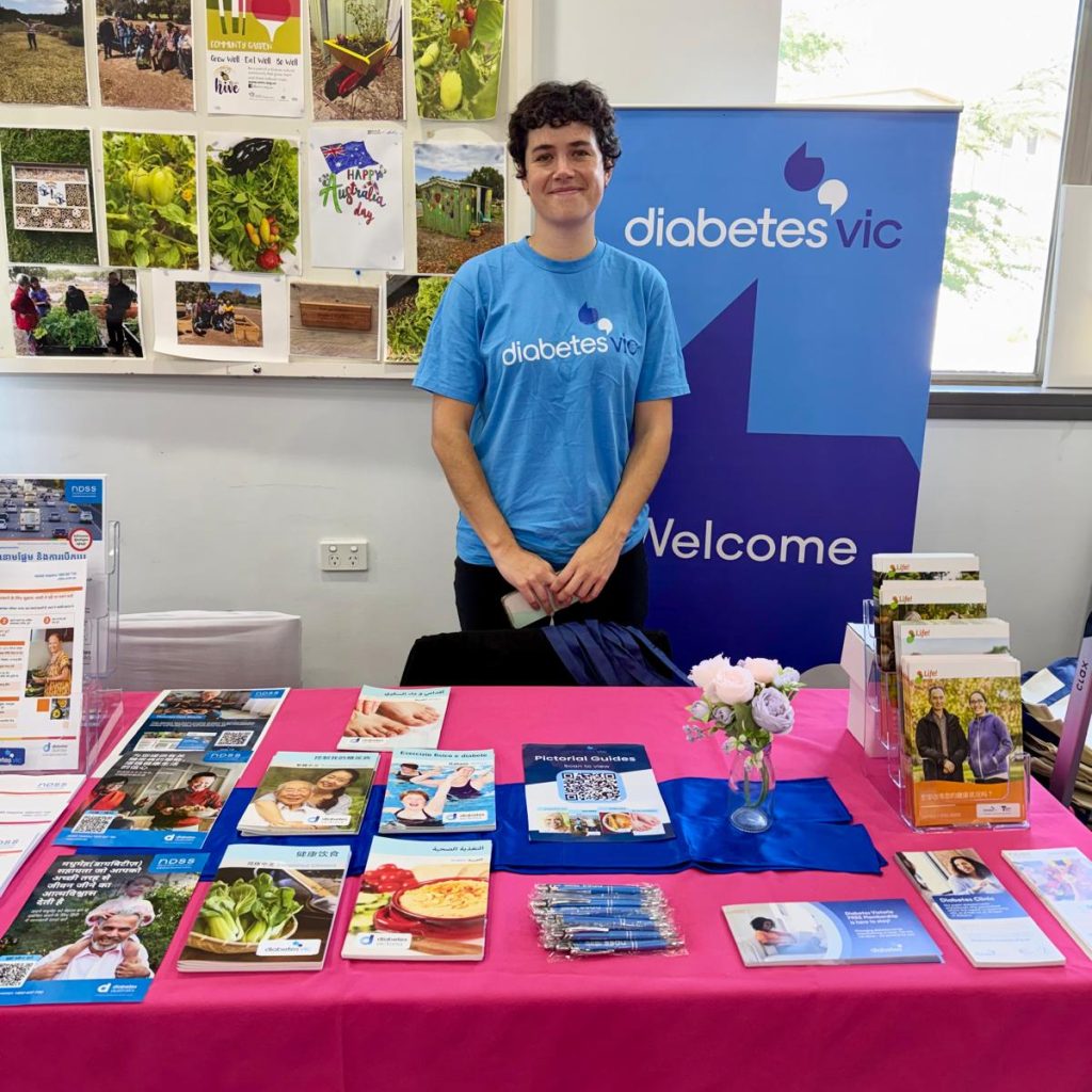A woman wearing a 'Diabetes Vic' blue t-shirt stands in behind a table of information. There is a Diabetes Vic banner behind them