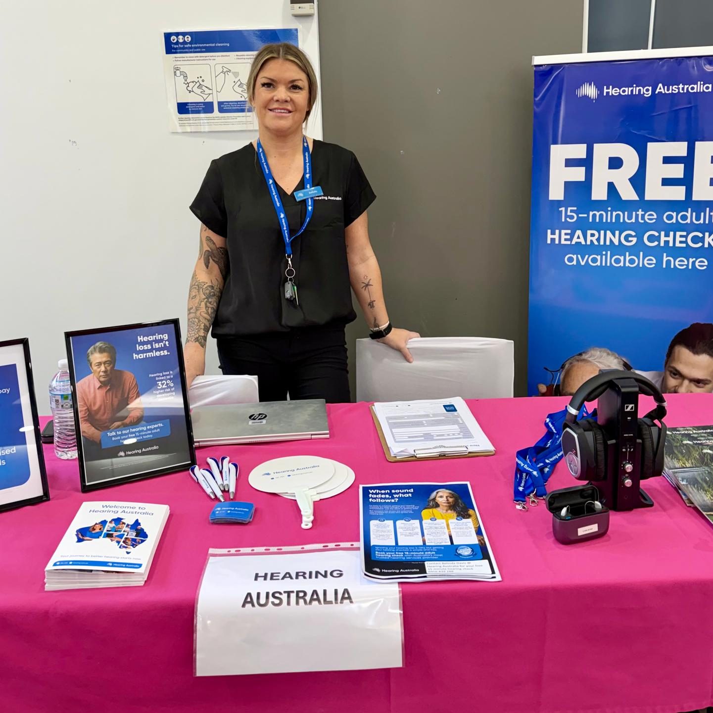 A woman stands behind a table of information from Hearing Australia. There is a blue banner next to them that says 'FREE 15-minute adult hearing checks available here