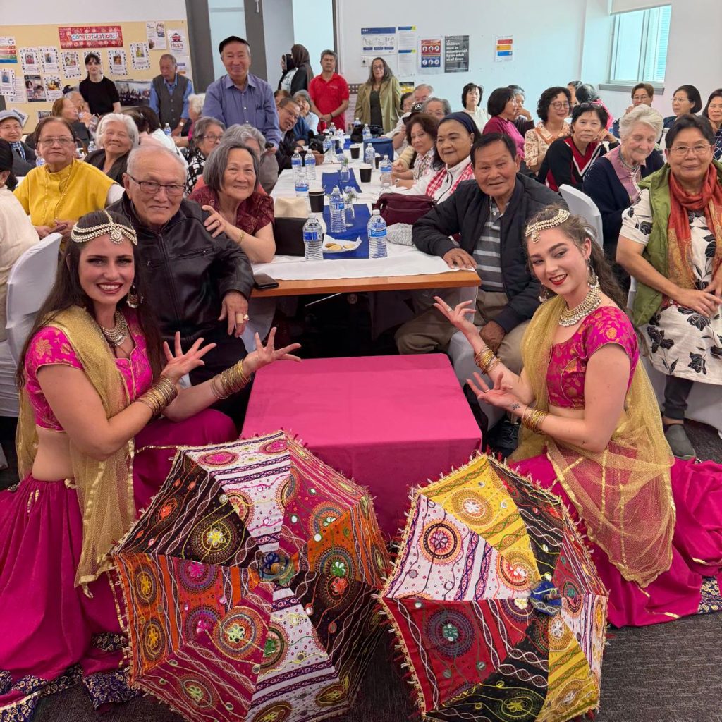 A pair of women dressed in bright cultural clothings kneels in front of a row of older people. They all smile at the camera.